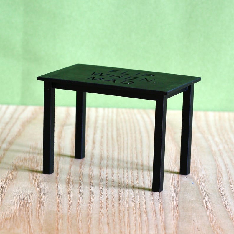 May include: A small black rectangular table with four thin legs. The top surface of the table features the embossed text "FLIP WHEN MAD" in a simple sans-serif font. The table has a smooth, matte finish and a minimalist, functional structure.