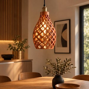May include: A terracotta-coloured pendant light with a teardrop shape and a lattice cutout pattern. The fixture features a brass-toned metal socket at the top, suspended by a black cord, and emits a warm glow from an internal light bulb.