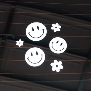May include: White vinyl stickers featuring smiley faces and flower shapes. The stickers are arranged in a playful pattern, perfect for decorating cars, laptops, or other surfaces. The design is simple and cheerful.