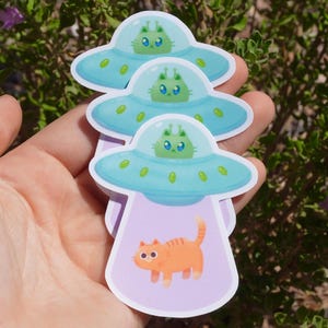 May include: Three stickers featuring a light blue UFO with a green alien inside. The bottom sticker shows an orange cat being beamed up. The stickers are held in a hand against a background of green leaves.