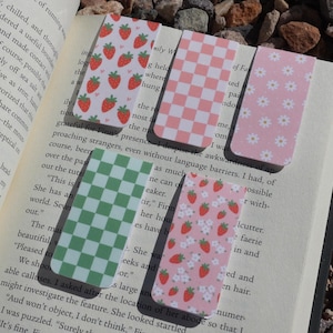 May include: A collection of colorful rectangular bookmarks with various patterns, including strawberries, daisies, and checkered designs. The bookmarks are placed on the pages of an open book.