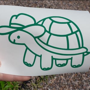 May include: A white rectangular decal featuring a dark green line art illustration of a turtle wearing a cowboy hat. The turtle is depicted in profile facing left, with a simple shell design and four legs visible on a plain white background.