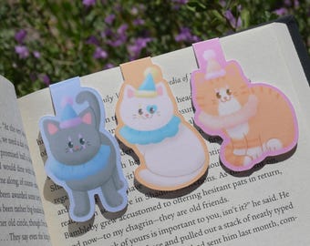 May include: Three cat-shaped bookmarks with party hats and ruffs are placed on the pages of an open book. The bookmarks are gray, white, and orange, with blue, pink, and yellow accents. The book is open to a page of printed text.