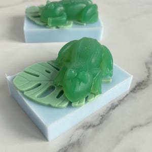 May include: Two handmade soaps, each featuring a green frog on a light green leaf, atop a light blue rectangular base. The soaps are likely scented and decorative, perfect for a bathroom or as a gift.