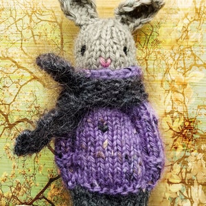 May include: A knitted gray bunny wearing a purple sweater and a black scarf. The bunny is standing against a background of a blurry image of a tree with yellow leaves.