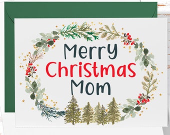 May include: A white Christmas card with a green envelope. The card features a watercolor wreath with gold stars, green leaves, and red berries. The text "Merry Christmas Mom" is written in red and blue lettering. The card has a blank space for a message.