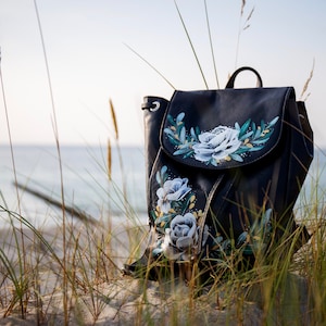 May include: A black leather backpack with a floral design painted on the front flap. The flowers are white with green leaves and yellow accents. The backpack is sitting on a sandy beach with tall grass in the background.