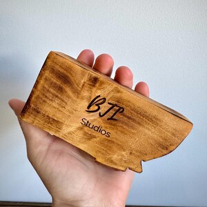 May include: A rectangular piece of light brown wood with a natural grain pattern, featuring the black text BTL Studios engraved on the front surface. The wood has a smooth, polished finish with darker, burnt-looking edges along the perimeter.