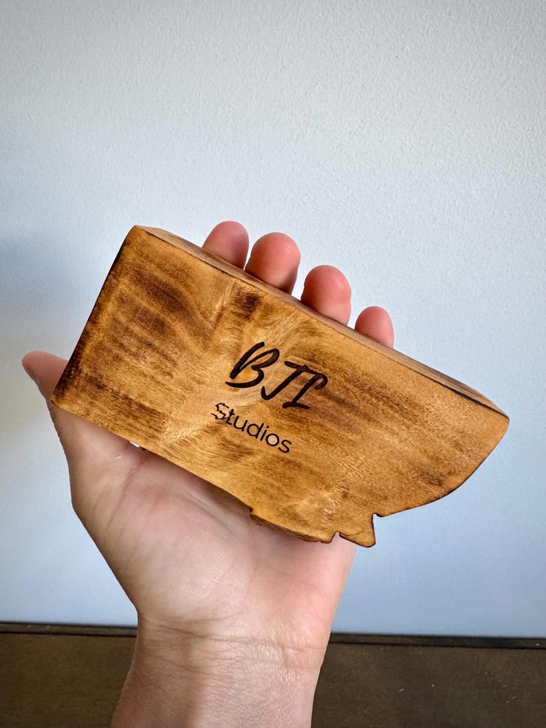 May include: A rectangular piece of light brown wood with a natural grain pattern, featuring the black text BTL Studios engraved on the front surface. The wood has a smooth, polished finish with darker, burnt-looking edges along the perimeter.