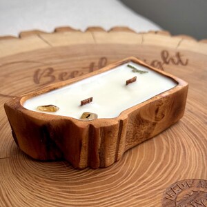 May include: A rectangular candle in a natural wood bowl with a light-colored wax fill. The candle features two wooden wicks and two small, smooth, amber-colored stones embedded in the wax. The wood bowl has a rough, textured bark-like exterior.