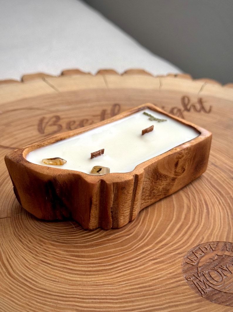 May include: A rectangular candle in a natural wood bowl with a light-colored wax fill. The candle features two wooden wicks and two small, smooth, amber-colored stones embedded in the wax. The wood bowl has a rough, textured bark-like exterior.