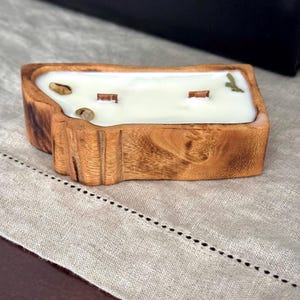 May include: A rectangular candle with a natural wood exterior and a white wax interior. The candle features two wooden wicks and is decorated with small, light brown stones and a green leaf embedded in the surface of the wax.