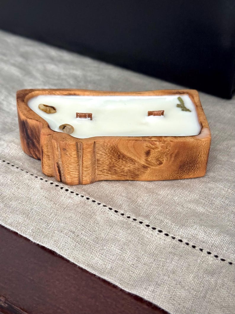May include: A rectangular candle with a natural wood exterior and a white wax interior. The candle features two wooden wicks and is decorated with small, light brown stones and a green leaf embedded in the surface of the wax.