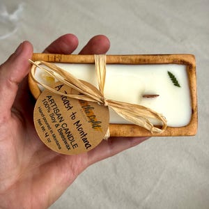 May include: A rectangular wooden dough bowl candle filled with white wax, a single wooden wick, and a small green leaf embedded on top. A round tan paper tag tied with raffia reads A Toast to Montana, Artisan Candle, 100% Soy & Beeswax, Net Wt: 4 oz.