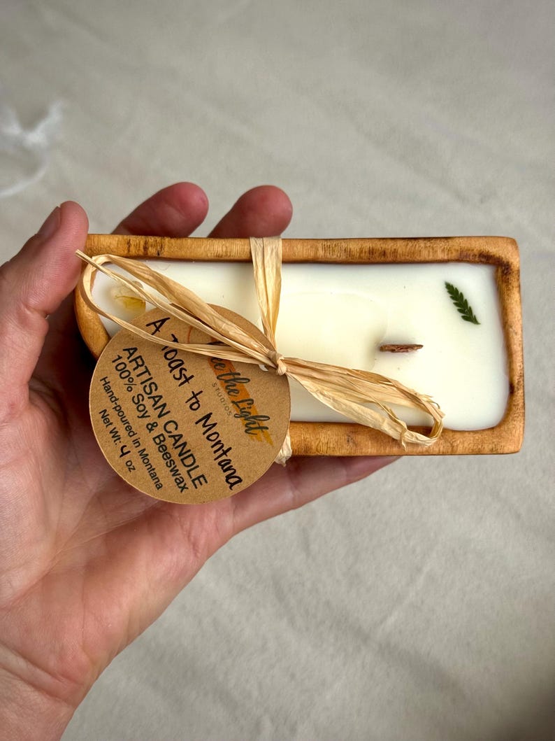 May include: A rectangular wooden dough bowl candle filled with white wax, a single wooden wick, and a small green leaf embedded on top. A round tan paper tag tied with raffia reads A Toast to Montana, Artisan Candle, 100% Soy & Beeswax, Net Wt: 4 oz.