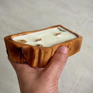 May include: A rectangular candle in a natural wood bowl with a light-colored wax surface. The candle features three wooden wicks and is decorated with small, smooth, light-brown stones embedded in the wax near the edges of the container.