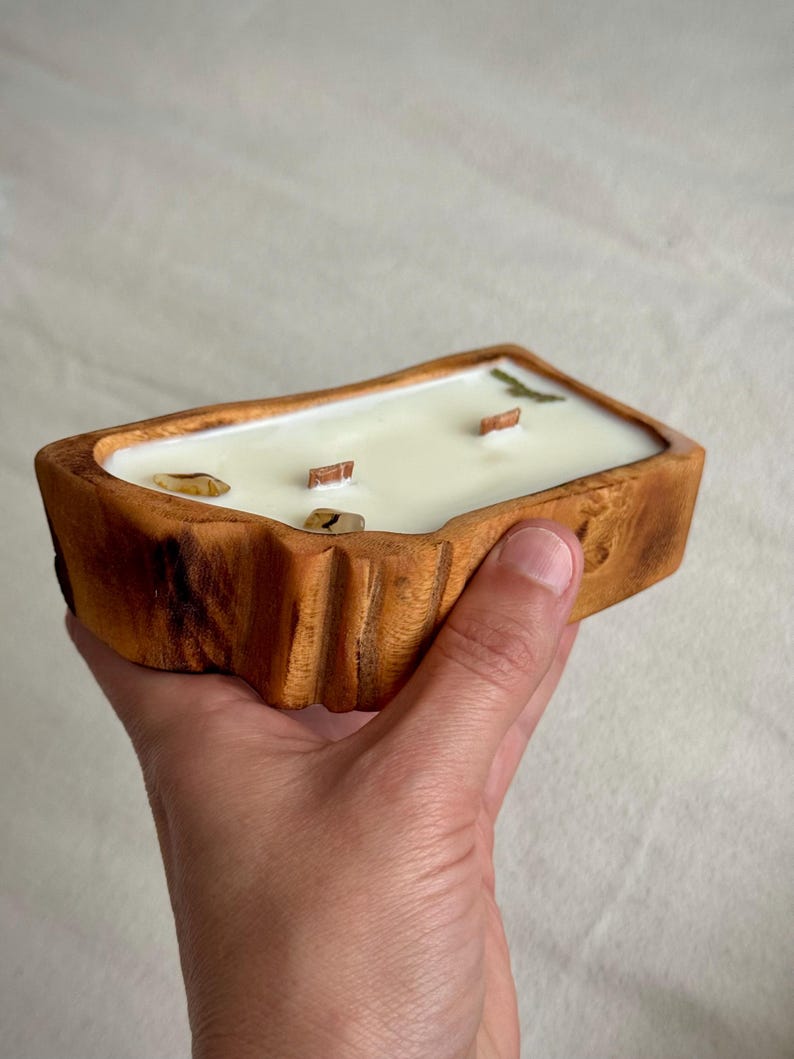 May include: A rectangular candle in a natural wood bowl with a light-colored wax surface. The candle features three wooden wicks and is decorated with small, smooth, light-brown stones embedded in the wax near the edges of the container.