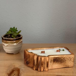 May include: A rectangular wooden candle container with a natural grain finish, filled with white wax and featuring two wooden wicks. The container has a carved, textured side detail and is positioned next to a small succulent in a ceramic pot.