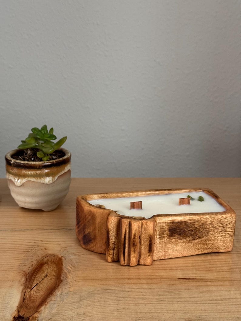 May include: A rectangular wooden candle container with a natural grain finish, filled with white wax and featuring two wooden wicks. The container has a carved, textured side detail and is positioned next to a small succulent in a ceramic pot.