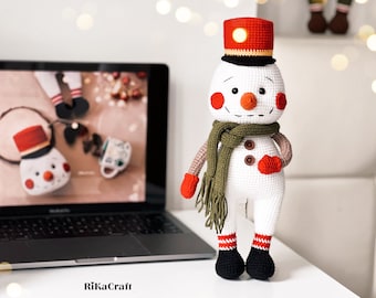 Peut inclure: Un bonhomme de neige en crochet blanc avec un chapeau rouge, des yeux noirs et une écharpe verte. Le bonhomme de neige tient une moufle rouge dans une main.