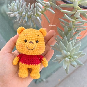 Pu&ograve; includere: Un peluche Winnie the Pooh all'uncinetto con un corpo giallo, una maglietta rossa e un naso marrone. Il giocattolo &egrave; seduto nella mano di una persona.