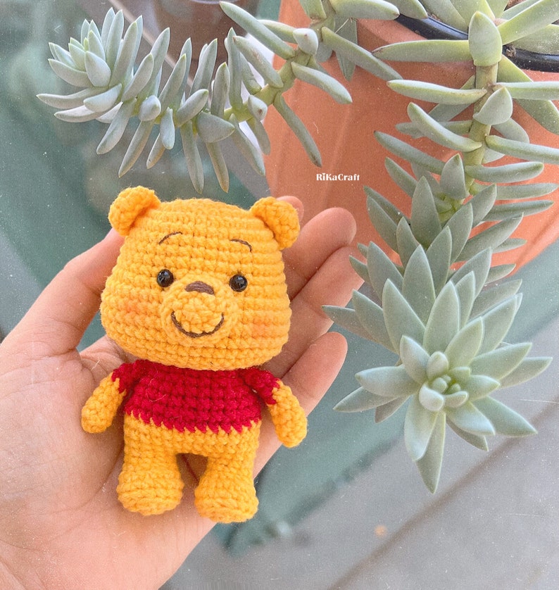 Pu&ograve; includere: Un peluche Winnie the Pooh all'uncinetto con un corpo giallo, una maglietta rossa e un naso marrone. Il giocattolo &egrave; seduto nella mano di una persona.