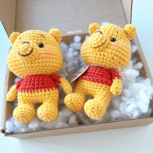 Pu&ograve; includere: Due peluche Winnie the Pooh all'uncinetto in una scatola con noccioline di imballaggio bianche. I giocattoli sono gialli con camicie rosse e hanno occhi e nasi neri.
