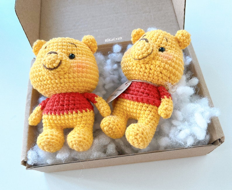 Pu&ograve; includere: Due peluche Winnie the Pooh all'uncinetto in una scatola con noccioline di imballaggio bianche. I giocattoli sono gialli con camicie rosse e hanno occhi e nasi neri.