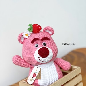 May include: A handmade pink crochet bear with a red nose and a strawberry on its head. The bear has a white belly and is sitting in a wooden crate. A tag with a floral design hangs from the bear.