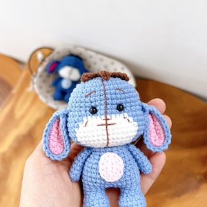 Puede incluir: Un juguete de burro de ganchillo azul hecho a mano con orejas internas rosas y un hocico blanco. El juguete tiene un mechón de pelo marrón y un círculo rosa en la barriga. Otro juguete similar está en el fondo.