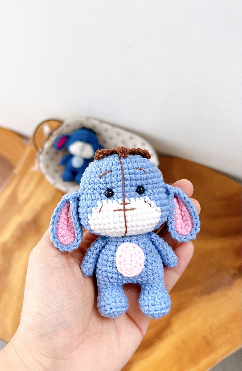 K&ouml;nnte beinhalten: Ein handgefertigtes blaues H&auml;keltier in Form eines Esels mit rosa Innenohren und einer wei&szlig;en Schnauze. Das Spielzeug hat einen braunen Haarschopf und einen rosa Kreis auf dem Bauch. Ein weiteres &auml;hnliches Spielzeug ist im Hintergrund.
