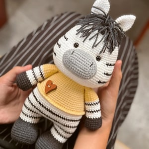 Puede incluir: Un peluche de cebra hecho a mano con ganchillo. La cebra tiene un cuerpo blanco con rayas negras, pezuñas grises y una camisa amarilla con un corazón de madera. La melena y la cola de la cebra son de color gris oscuro.