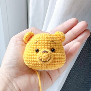 Pu&ograve; includere: Un personaggio di Winnie the Pooh all'uncinetto, realizzato con filato giallo e occhi neri. Il personaggio tiene un piccolo naso marrone.