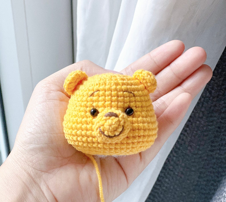 Pu&ograve; includere: Un personaggio di Winnie the Pooh all'uncinetto, realizzato con filato giallo e occhi neri. Il personaggio tiene un piccolo naso marrone.