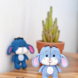 K&ouml;nnte beinhalten: Zwei handgefertigte Eeyore-Figuren aus H&auml;kelarbeit in Hellblau mit rosa Innenohren und einem wei&szlig;en Bauch. Eine ist scharf, die andere im Hintergrund verschwommen. Eine kleine Topfpflanze steht dahinter auf einer Holzoberfl&auml;che. Die Figuren sind etwa 10 cm gro&szlig;.