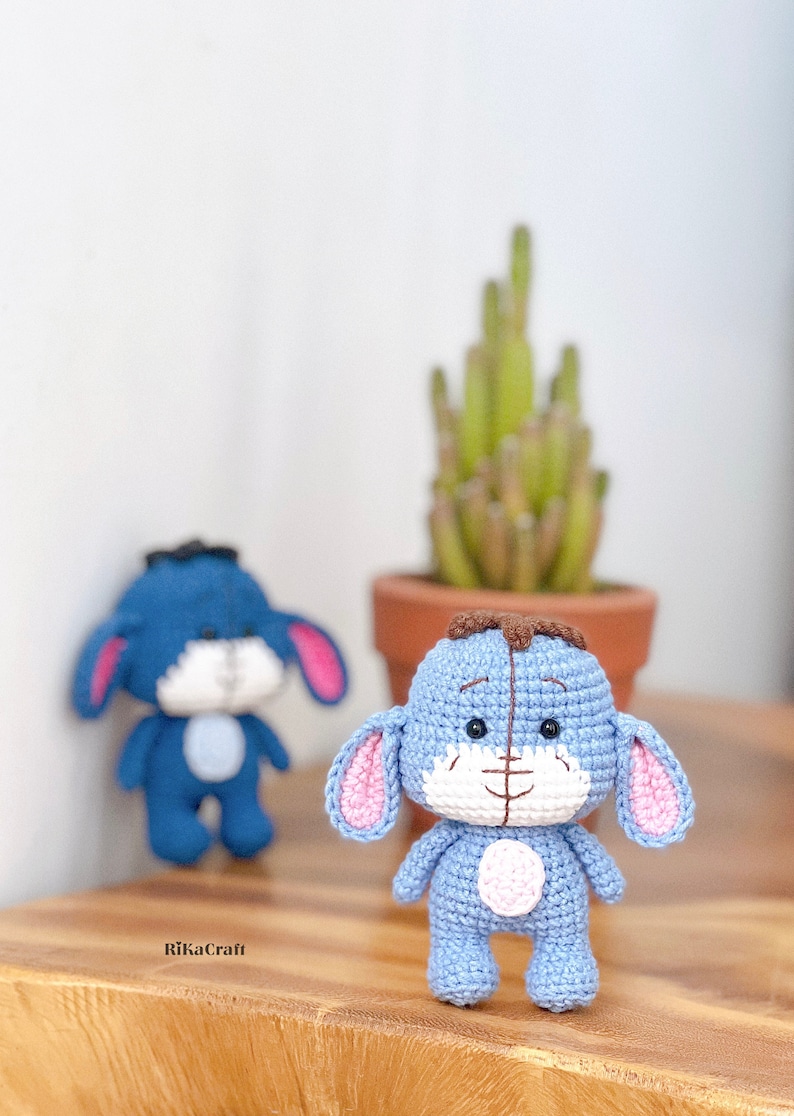 K&ouml;nnte beinhalten: Zwei handgefertigte Eeyore-Figuren aus H&auml;kelarbeit in Hellblau mit rosa Innenohren und einem wei&szlig;en Bauch. Eine ist scharf, die andere im Hintergrund verschwommen. Eine kleine Topfpflanze steht dahinter auf einer Holzoberfl&auml;che. Die Figuren sind etwa 10 cm gro&szlig;.