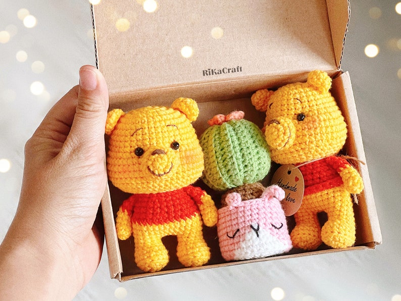 Pu&ograve; includere: Una scatola di cartone marrone con un orsacchiotto Winnie the Pooh di uncinetto giallo, un cactus di uncinetto verde e un vaso di uncinetto rosa con un viso bianco. La scatola ha il testo "RikaCraft" stampato sulla parte superiore e un'etichetta che dice "Fatto a mano con amore".