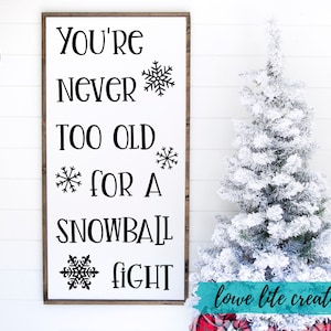 May include: A white sign with black text that reads "You're never too old for a snowball fight."  The sign is decorated with snowflakes. The sign is hanging on a white wall with a snowy Christmas tree in the background.