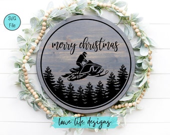 May include: A round wooden sign with a gray background and black text that reads "merry christmas". A black silhouette of a snowmobile driving through a forest is in the center of the sign. The sign is surrounded by a garland of green leaves and brown beads.