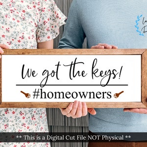 Pode incluir: Um casal segura uma placa de madeira com o texto "We got the keys! #homeowners" em escrita preta. A placa tem dois ícones de chaves marrons de cada lado do texto. O casal está vestindo roupas casuais.