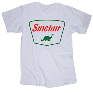 May include: White t-shirt with a green and red Sinclair logo featuring a green dinosaur. The logo is in a shield shape.