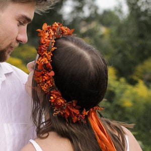 Può includere: Una donna con lunghi capelli castani indossa una corona di fiori fatta di fiori arancioni e marroni con nastri arancioni. La corona è indossata sulla parte superiore della testa e i nastri pendono sulla schiena.