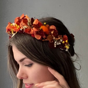 Può includere: Una donna indossa un abito di pizzo grigio con una corona di fiori fatta di fiori secchi in tonalità arancio e rosso.