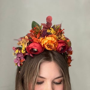 Può includere: Un cerchietto floreale con fiori arancioni, rossi e gialli. Il cerchietto è fatto di fiori e verde artificiali.
