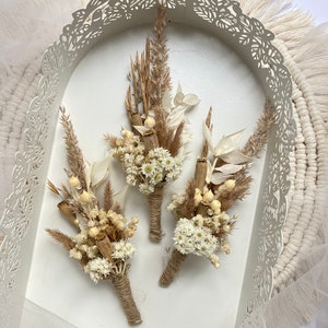 May include: Three small bouquets of dried flowers, each tied with twine. The bouquets are made with white and brown dried flowers and grasses. The bouquets are arranged in a white tray with a decorative cut-out design.