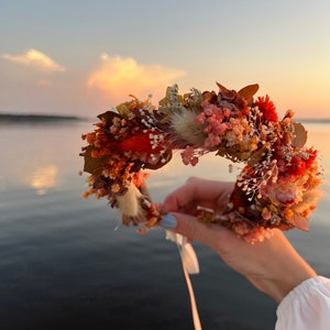Può includere: Una corona di fiori secchi con un mix di fiori rossi, arancioni e rosa. La corona è tenuta in una mano sullo sfondo di un lago e del cielo.