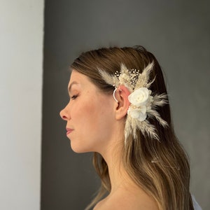 Puede incluir: Un pendiente de oreja floral blanco con flores secas y plumas. El pendiente de oreja se lleva en la oreja de una persona.
