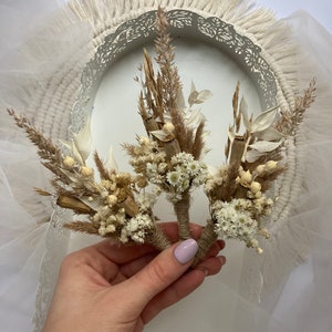 May include: Three small bouquets of dried flowers, tied with twine, in a hand. The bouquets are made of white and brown dried flowers, including small white flowers, dried grasses, and dried leaves.