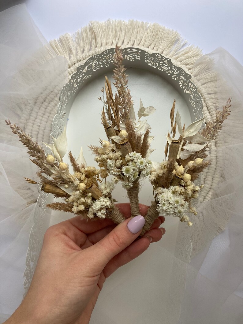 May include: Three small bouquets of dried flowers, tied with twine, in a hand. The bouquets are made of white and brown dried flowers, including small white flowers, dried grasses, and dried leaves.