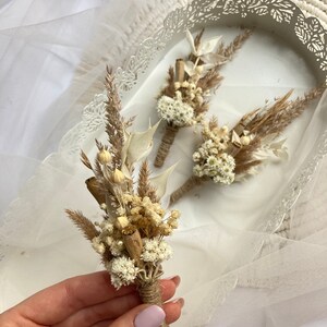 May include: Three small bouquets of dried flowers, tied with twine, in a white tray. The bouquets are made of white and beige dried flowers and grasses.